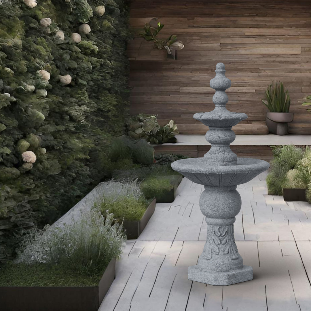 A Teamson Home Icy Stone 2-Tier Waterfall Garden Fountain, Gray, a durable statue, stands in an outdoor garden with wooden walls and lush greenery.