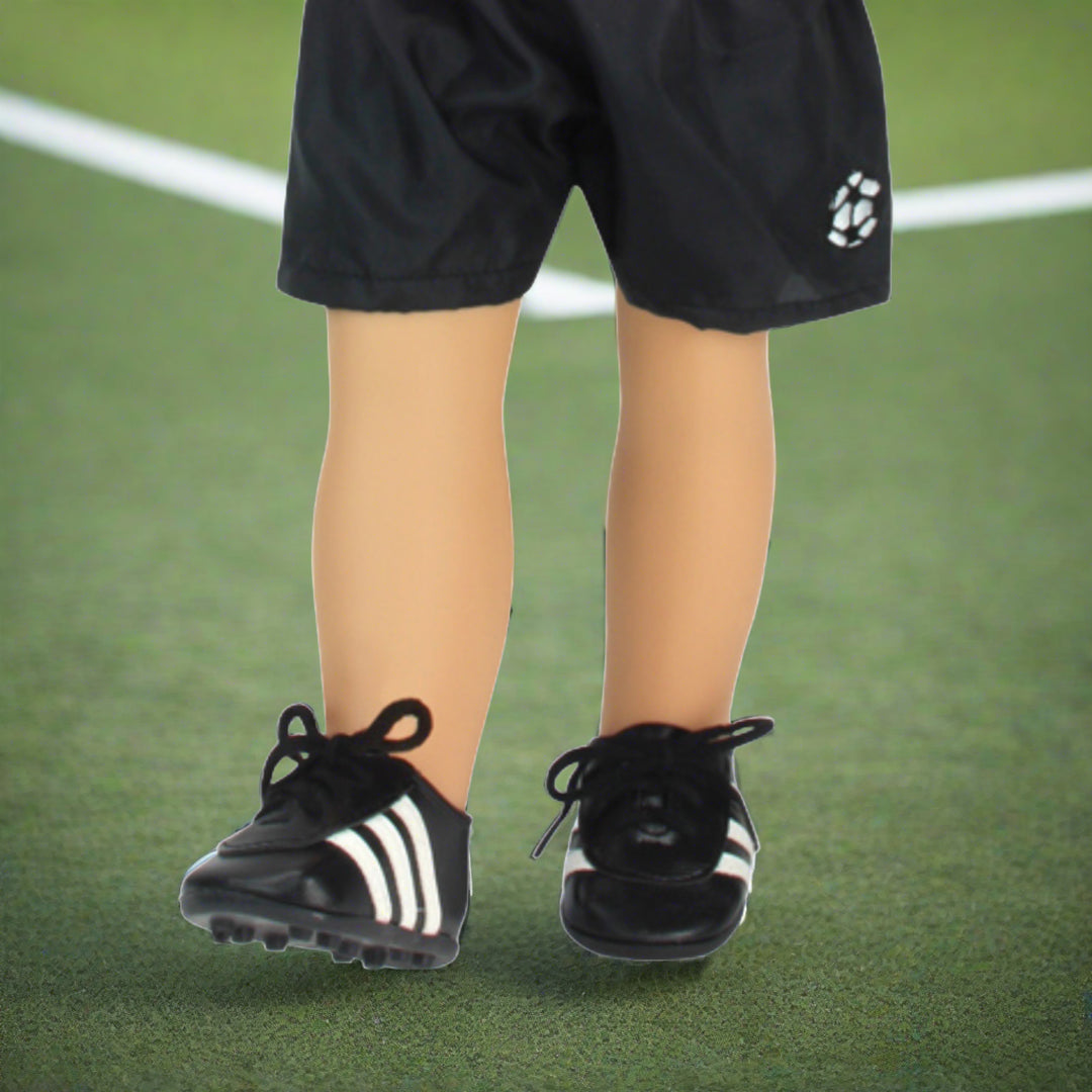 A close-up of a pair of black soccer shorts and cleats on an 18" doll standing on a pretend soccer field.