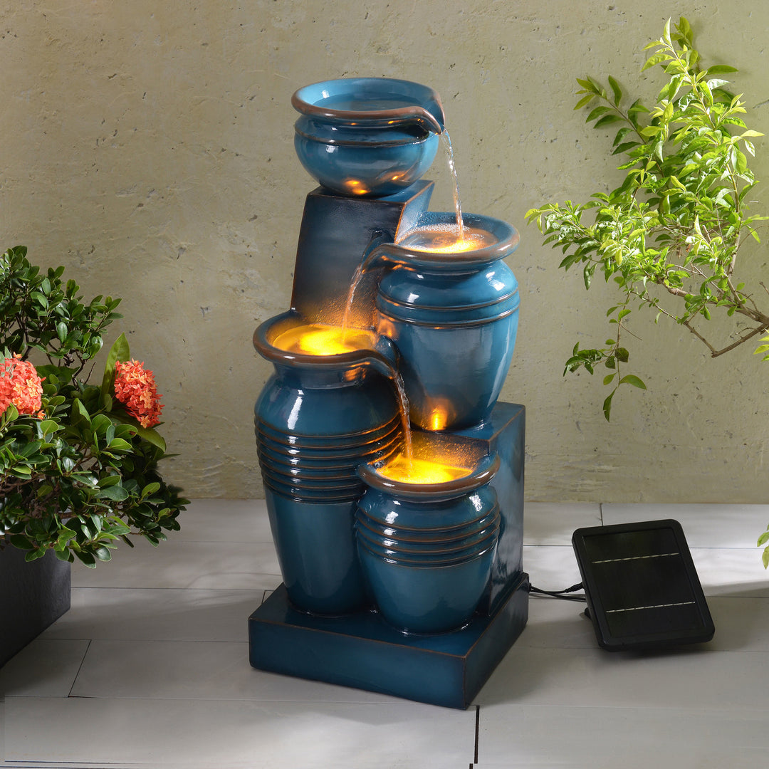Teamson Home 28.74" Blue 4-Tier Outdoor Solar Water Fountain with LED Lights glowing underneath the flowing water, with plants on either side, next to the solar panel