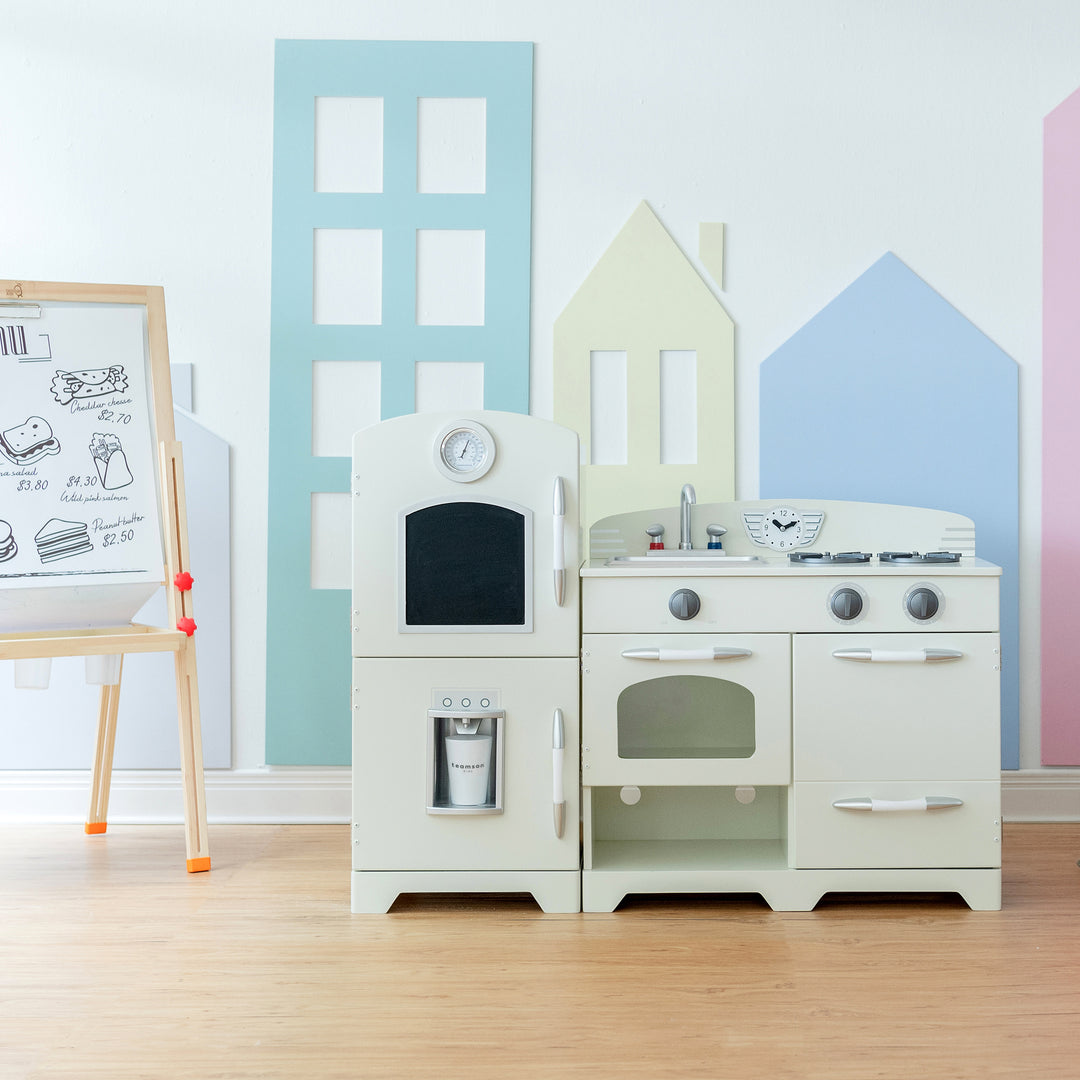 A children's playroom with an easy-to-clean Teamson Kids Little Chef Fairfield Retro Kids Kitchen Playset with Refrigerator, Ivory and easel, featuring pastel house silhouettes on the wall.