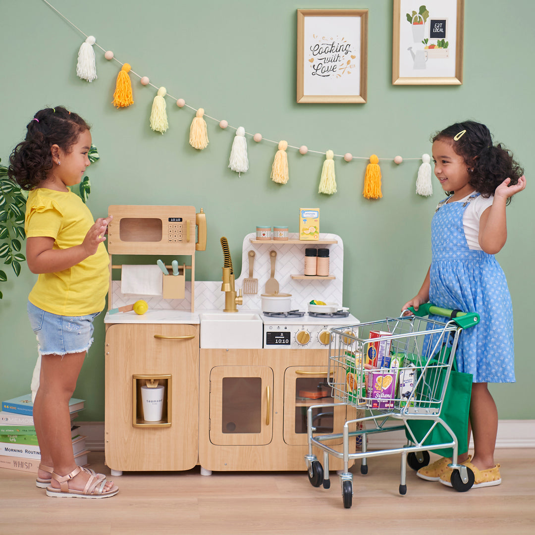 Two children playing with a Teamson Kids - Little Chef Cyprus Medium Play Kitchen, Light Oak/White and a shopping cart in a playroom.