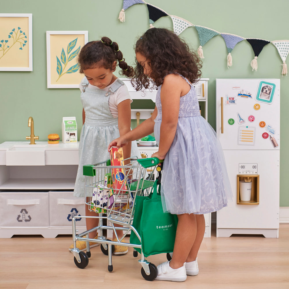 Two children play shopping with a Teamson Kids - Little Helper Dallas Shopping Cart with Play Food, Chrome/Green and groceries in a playroom with a kitchen set.