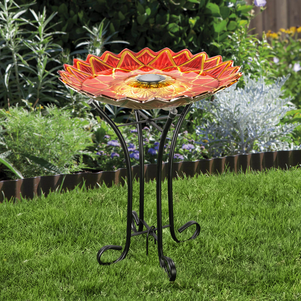A decorative 17.8" Dahlia Fusion Glass Birdbath with Solar-Powered Light in the shape of a red and yellow flower, mounted on a metal stand in a garden setting.