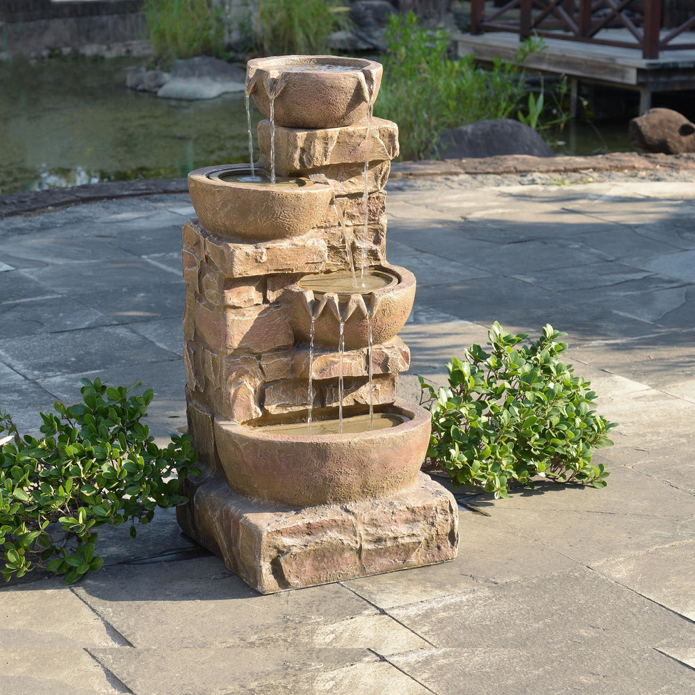 A 33.27" Cascading Bowls & Stacked Stones LED Outdoor Fountain with flowing water in a garden setting.