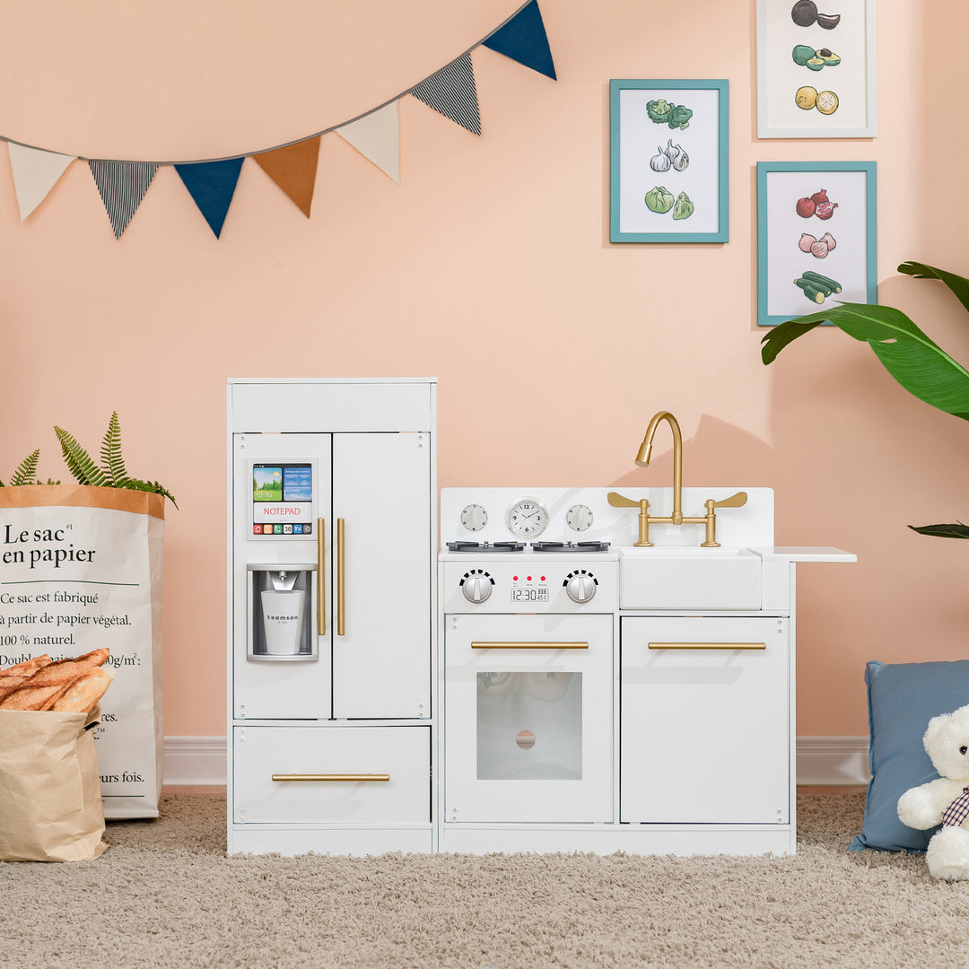 A child's Teamson Kids Little Chef Charlotte Modern Play Kitchen, White/Gold set in a room with wall decorations and a teddy bear.