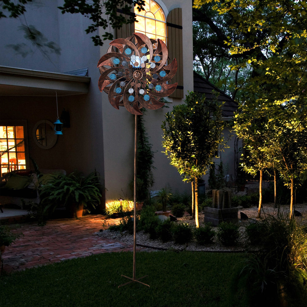 A 18" dia. x 70" H Solar Metallic Kinetic Windmill Spinner, Copper illuminated by a spotlight in a garden at dusk.
