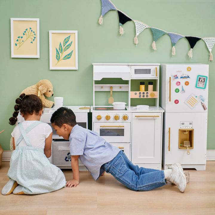 Two children playing with a Teamson Kids - Little Chef Atlanta Large Modular Play Kitchen, White/Gold in a room decorated with bunting.
