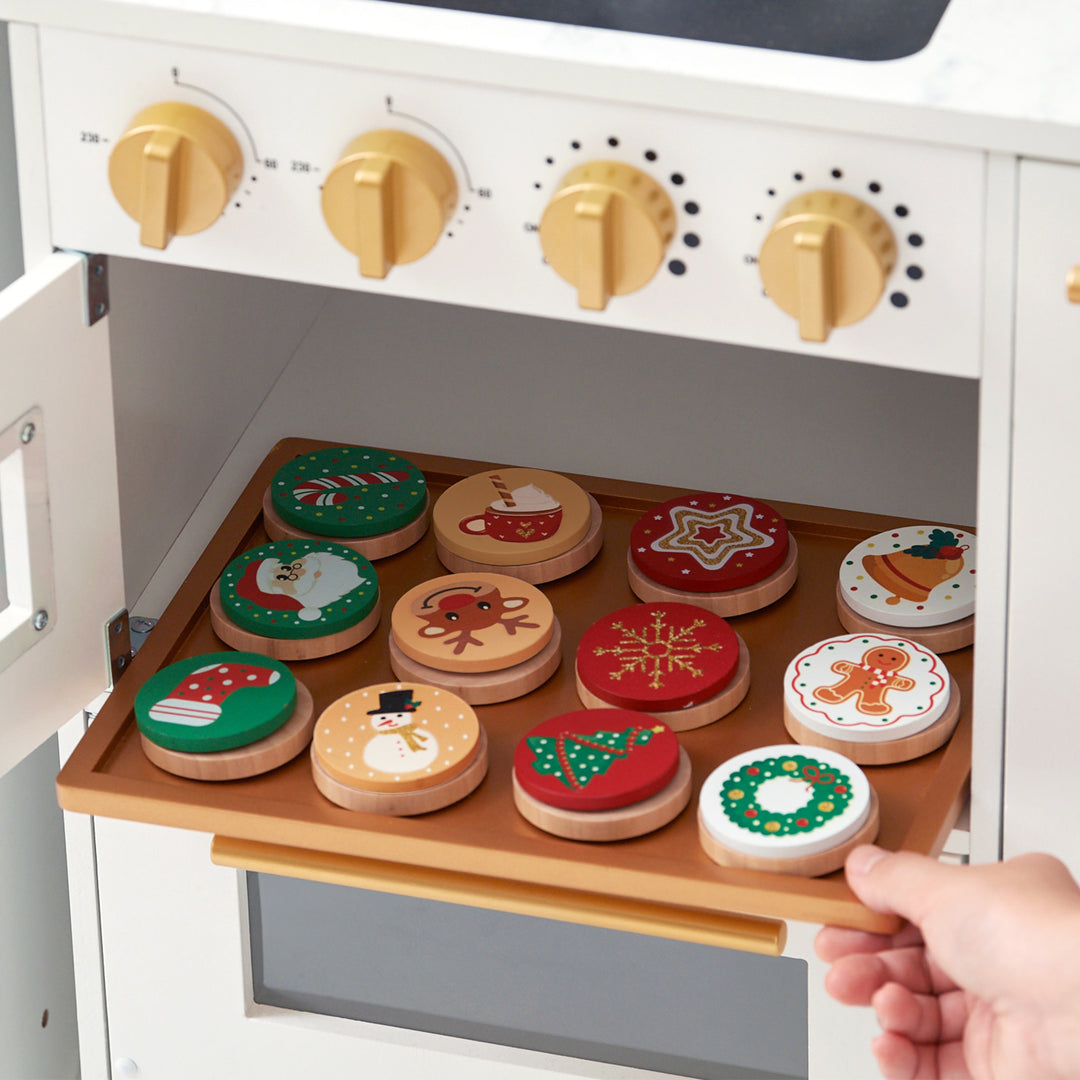 A child's hand placing a toy cookie into a Teamson Kids - Little Chef Atlanta Large Modular Play Kitchen, White/Gold filled with various decorated toy cookies.