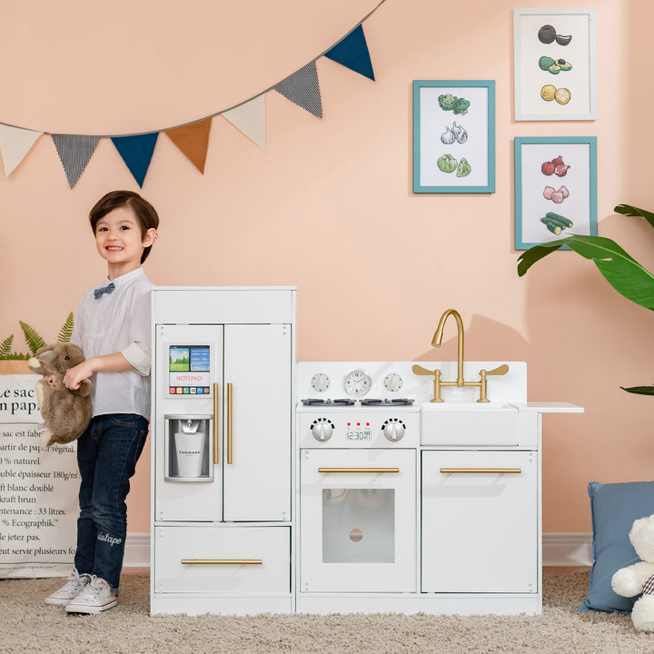 A child standing next to a Teamson Kids Little Chef Charlotte Modern Play Kitchen, White/Gold with interactive features in a playroom.