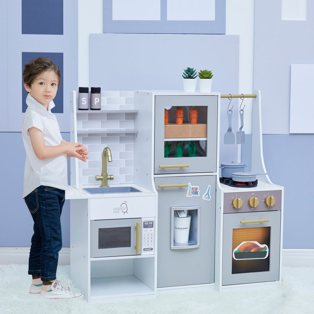 Boy standing in a grey room next to the Teamson Kids Little Chef Lyon Complete Wooden Kitchen Set with Hydroponic Garden, Refrigerator and Accessories, Gray