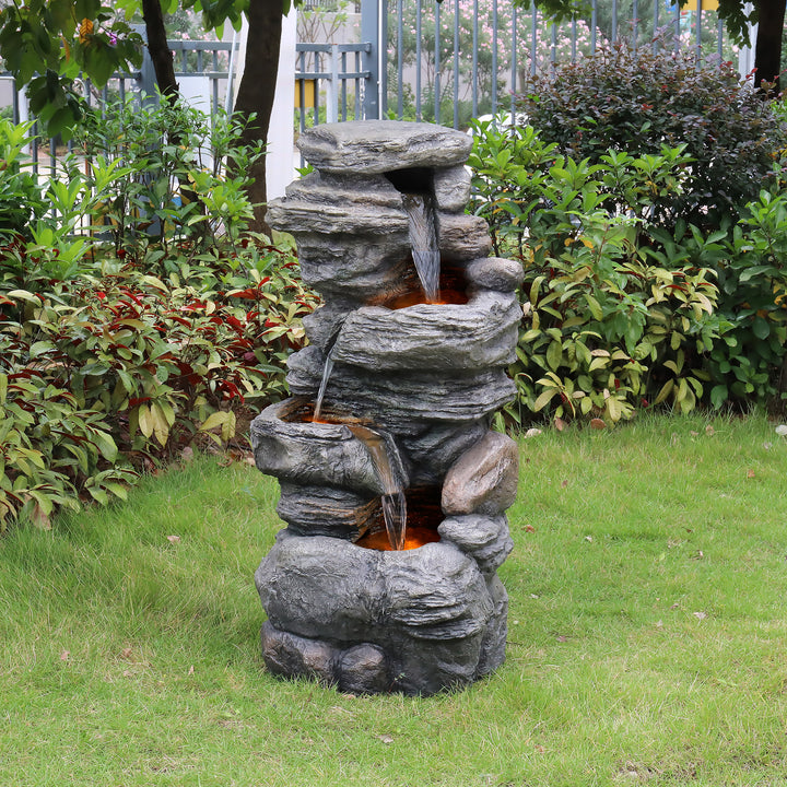 Teamson Home 4-Tier Stacked Stone Water Fountain with LED Lights, Gray, sat next to some shrubs in a yard