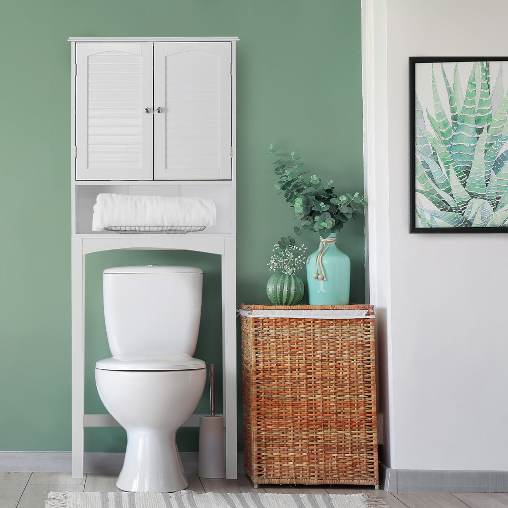 A tidy bathroom corner with a White Teamson Home Louis Over-the-Toilet Cabinet with Louvered Doors and an Open Shelf, a wicker laundry basket, against a green wall, decorated with plant-themed art and greenery.