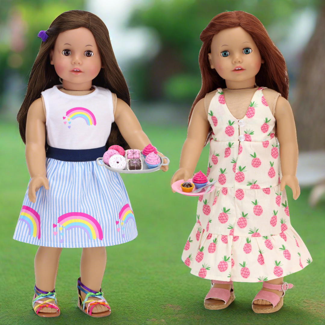 A brunette doll in a dress with rainbows and an auburn haired doll in a pink and ivory dress holding pretend baked goods.
