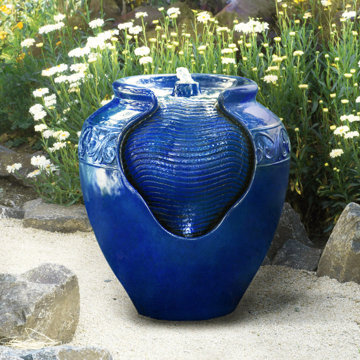 A Teamson Home Outdoor Glazed Pot Floor Fountain with LED Lights, Royal Blue surrounded by plants, enhanced with LED lights as garden decor.