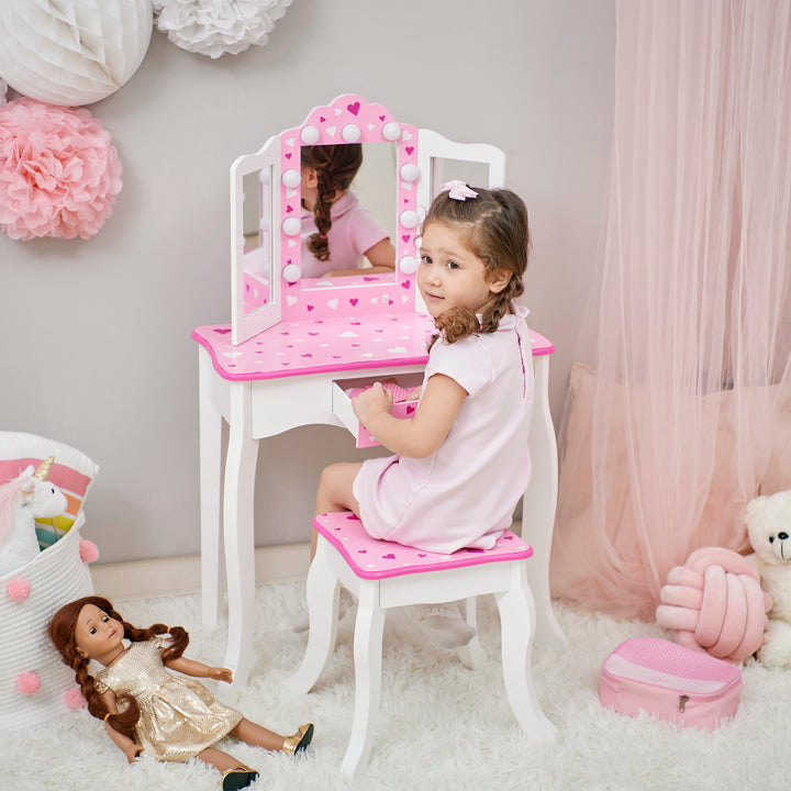 A little girl sits on a stool in front of white and pink vanity set with table and stool with pink heart accents and a lighted tri-fold mirror.