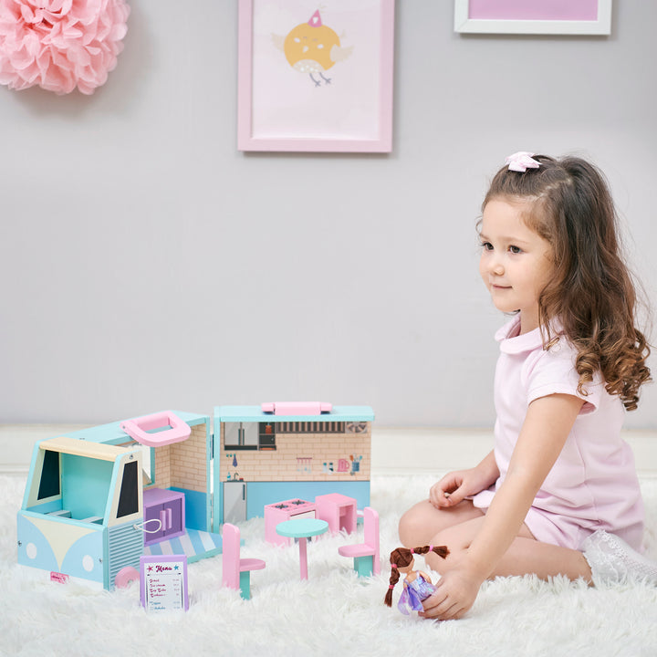 A girl is playing with a blue and pink food truck for dolls with a pink and white awning. The food truck is opened up so you can see the fully-illustrated interior.