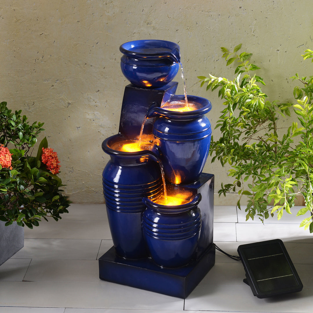 Teamson Home 28.74" Navy Blue 4-Tier Outdoor Solar Water Fountain with LED Lights on a patio with plants on either side