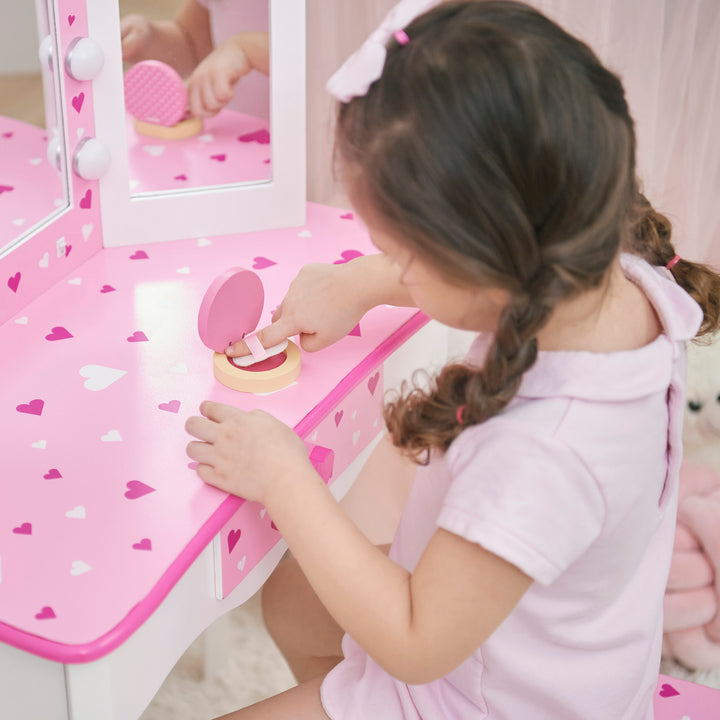 A little girl is playing white and pink vanity set with table and stool with pink heart accents and a lighted tri-fold mirror.