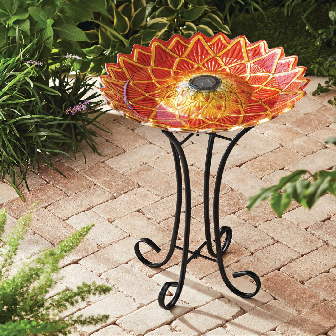 17.8" Dahlia Fusion Glass Birdbath with Solar-Powered Light, Red on a wrought iron stand, placed on a brick patio surrounded by greenery.