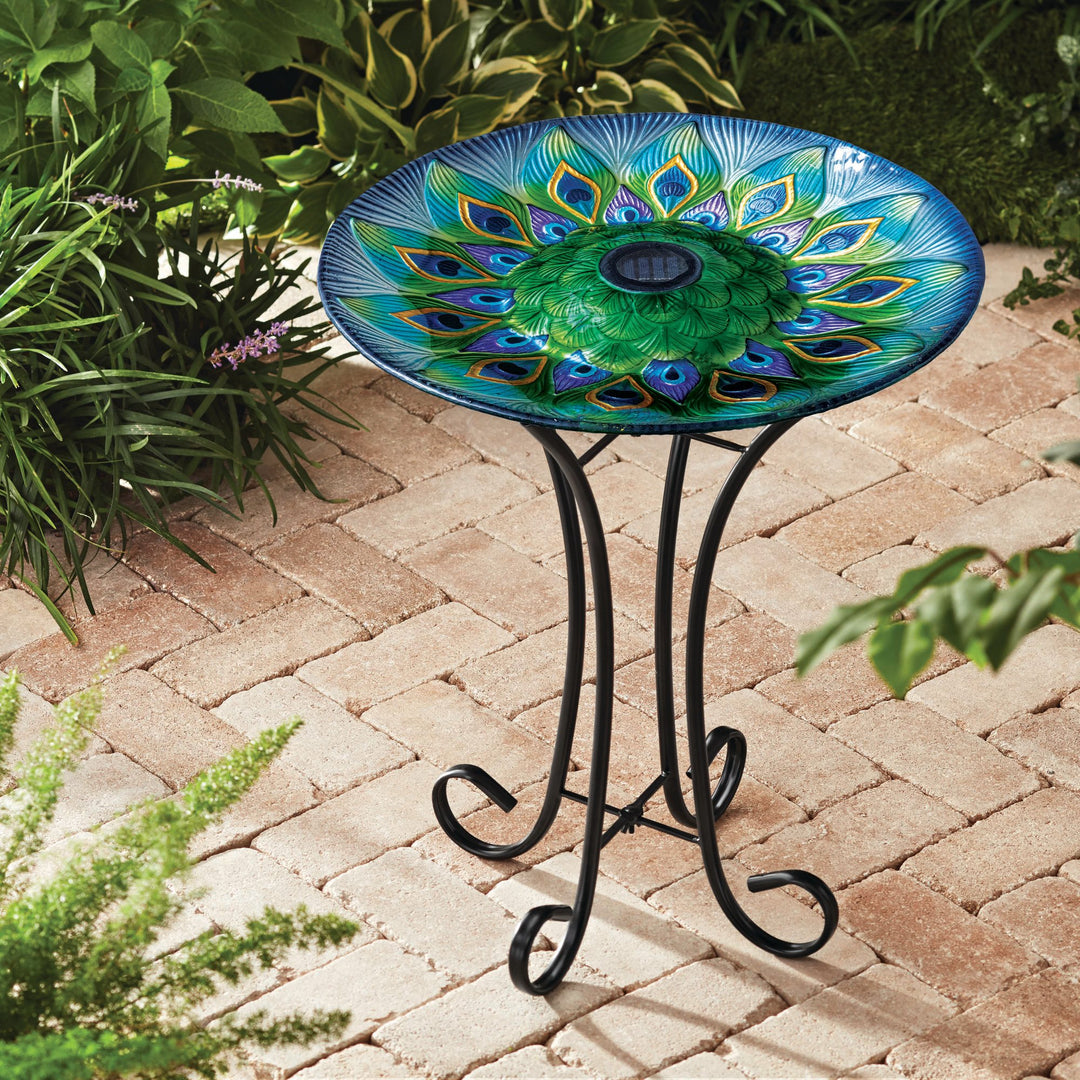 A 17.8" Peacock Fusion Glass Birdbath with Solar-Powered Light, Blue and Green, on a wrought iron stand set on a brick patio.
