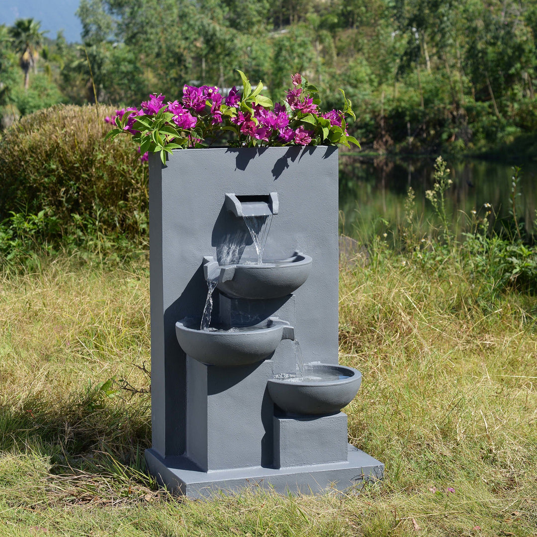 29.13" Outdoor Water Fountain with Planter & LED Lights, Matte Gray with purple flowers on top, set against a natural backdrop with a pond.