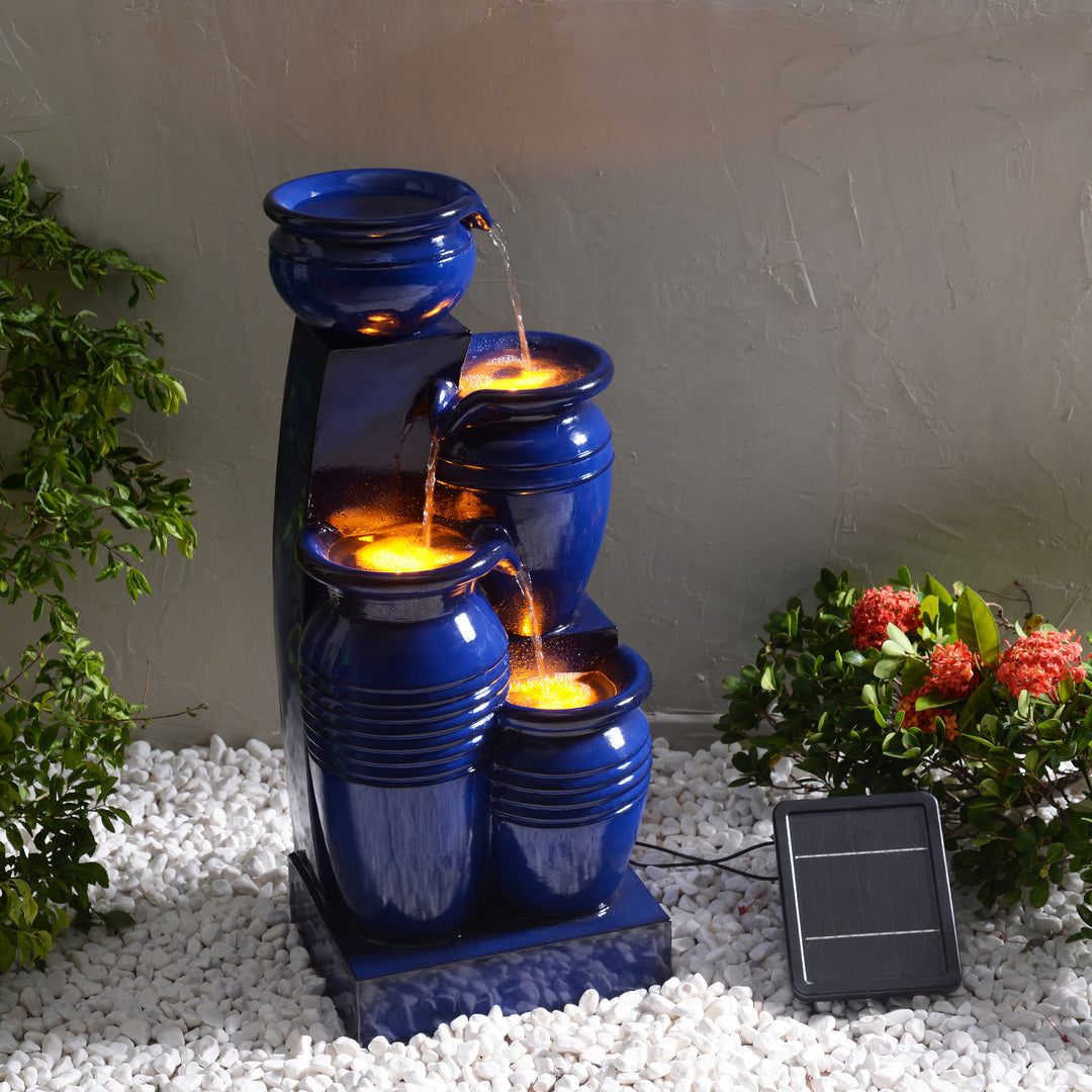 Teamson Home 28.74" Navy Blue 4-Tier Outdoor Solar Water Fountain with LED Lights alongside plants and white pebbles.