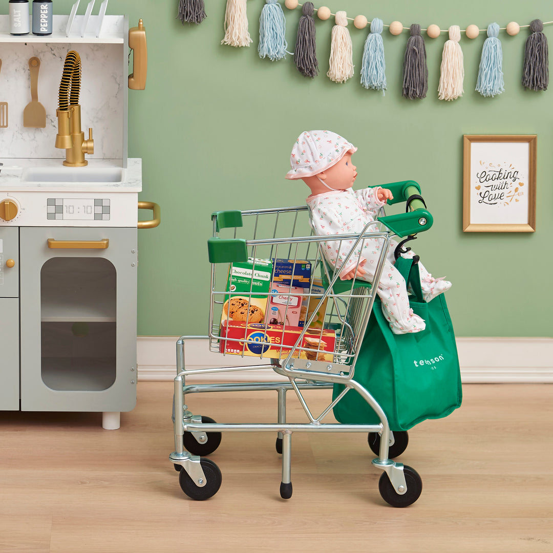 A baby doll sits in a Teamson Kids - Little Helper Dallas Shopping Cart with Play Food, Chrome/Green filled with pretend food items in a play kitchen setup.
