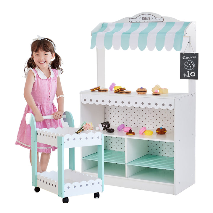 A young girl playing with a Teamson Kids My Dream Bakery Shop, Treat Stand and Dessert Cart, White/Blue filled with pretend pastries.