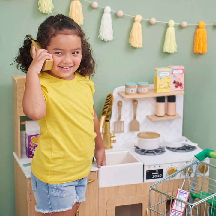 Young girl with a playful smile pretending to talk on a TEAMSON KIDS - LITTLE CHEF CYPRUS MEDIUM PLAY KITCHEN, LIGHT OAK/WHITE like a phone next to a toy kitchen set.