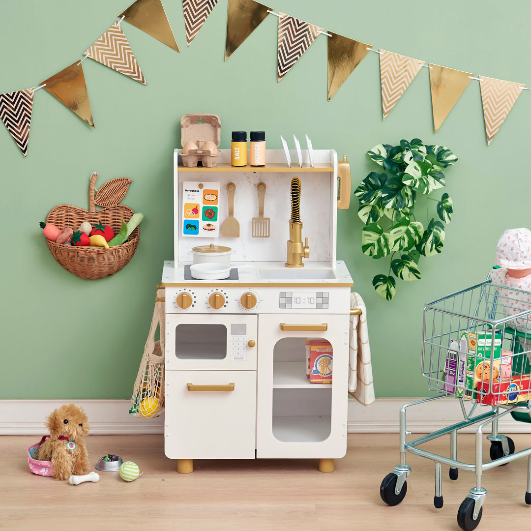 A kids play kitchen in white with gold accents placed against a green wall with accessories and a toy shopping cart in a room with green walls and decorative bunting.