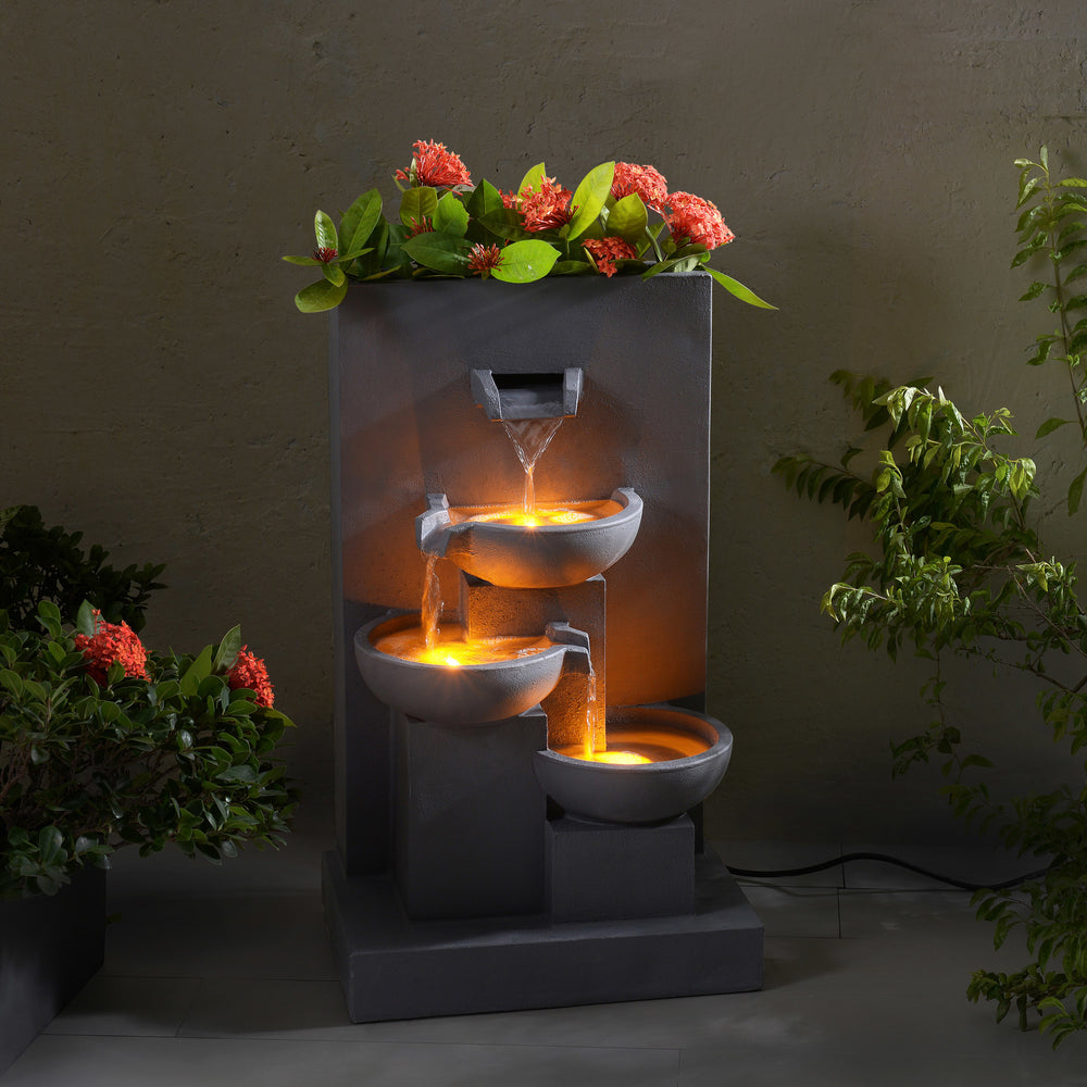 Outdoor tiered water fountain with ambient lighting and decorative plants in a evening setting
