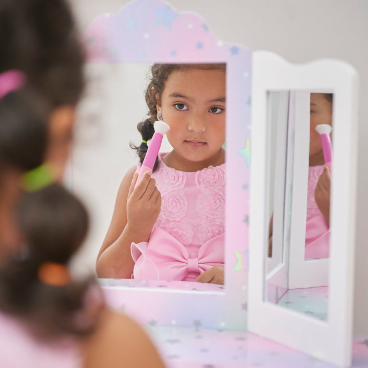 A little girl is brushing her hair in front of a mirror in the white vanity table with iridescent accents with silver stars.