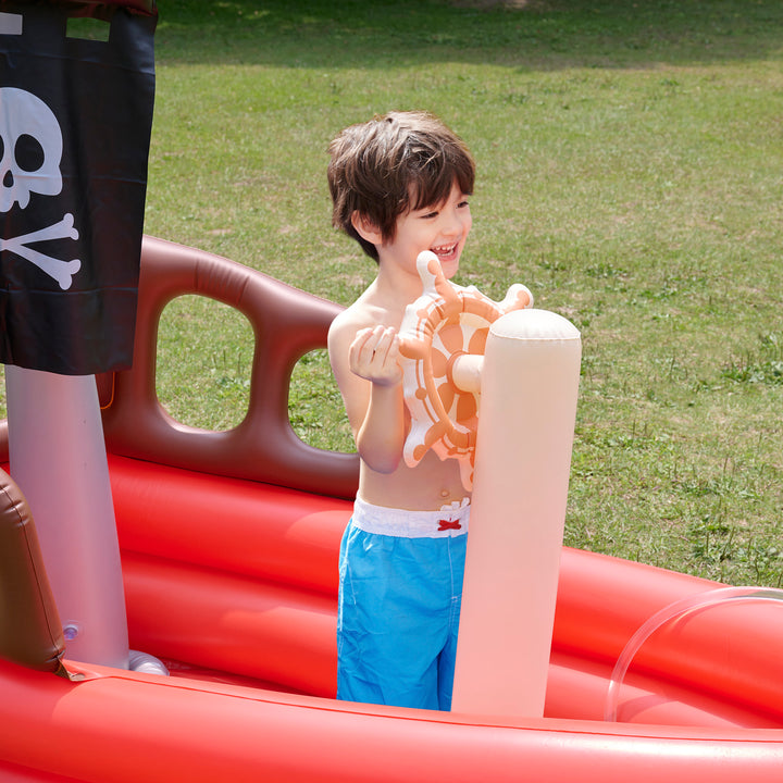 A child standing in the Teamson Kids - Water Fun Pirate boat pretending to turn the wheel, smiling.