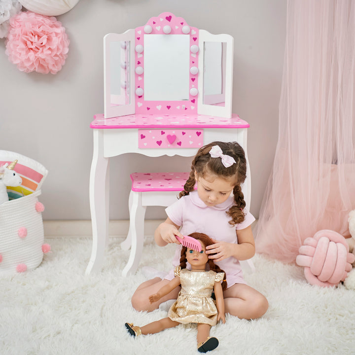 A little girl in a pink dress brushes her doll's hair sitting on the floor in front of a white and pink vanity set with table and stool with pink heart accents and a lighted tri-fold mirror.
