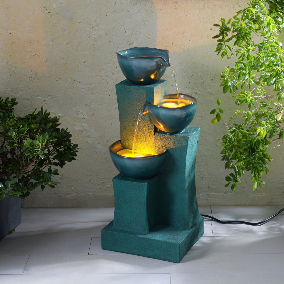 An illuminated 28.54" 3-Tier Outdoor Water Fountain with LED Lights, Green with water cascading through three bowl-shaped tiers, set next to a potted plant, against a textured backdrop.