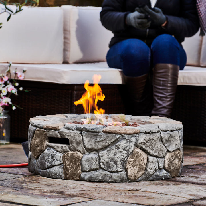 Person sitting by a Teamson Home 28" Outdoor Round Stone Propane Gas Fire Pit, Stone Gray with flames rising from the center.