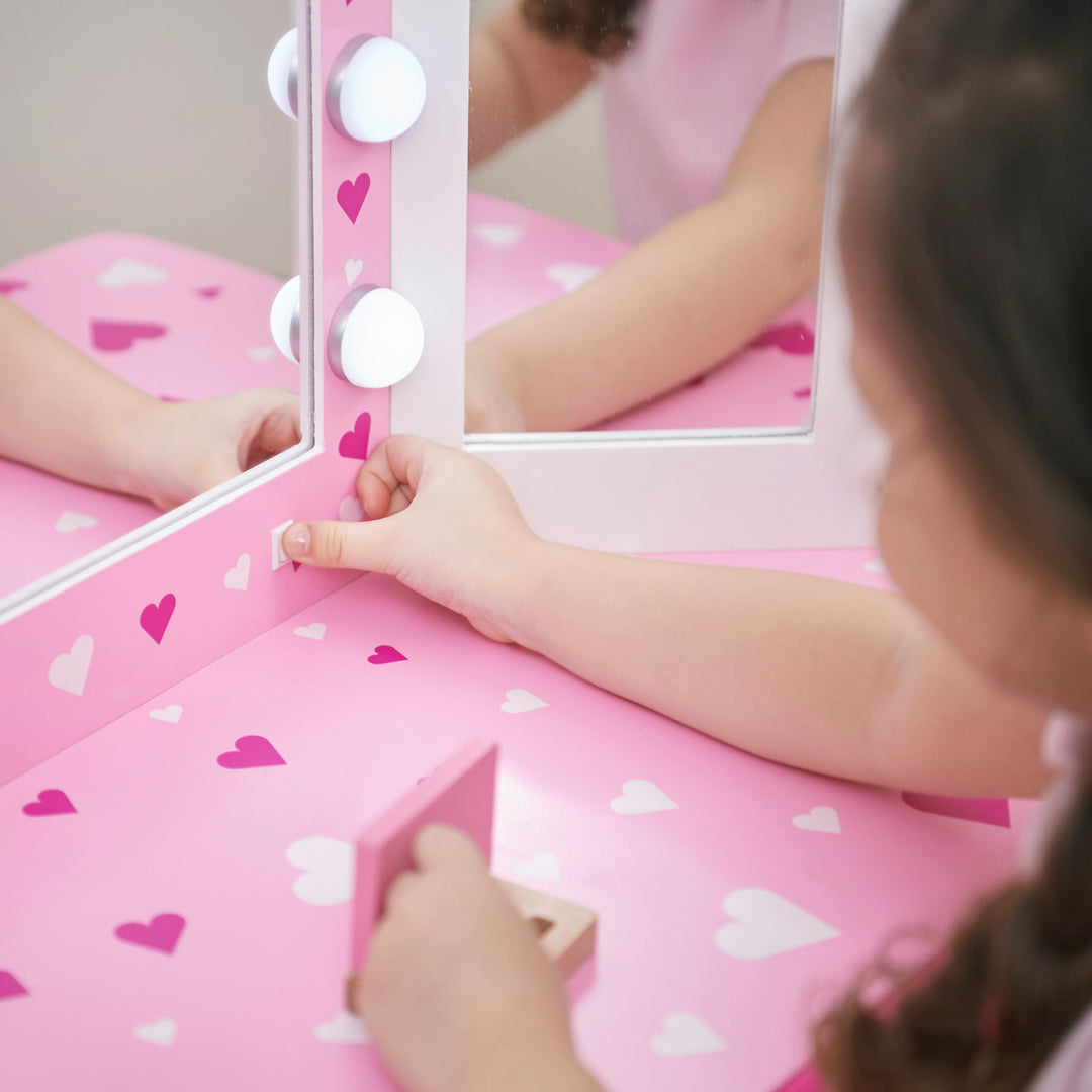A little girl is flicking the on/off white and pink vanity set with table and stool with pink heart accents and a lighted tri-fold mirror.