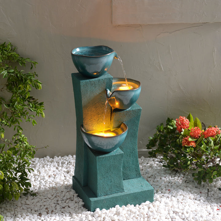 A 28.54" 3-Tier Outdoor Water Fountain with LED Lights, Green with cascading bowls positioned on white pebbles beside a flowering plant.