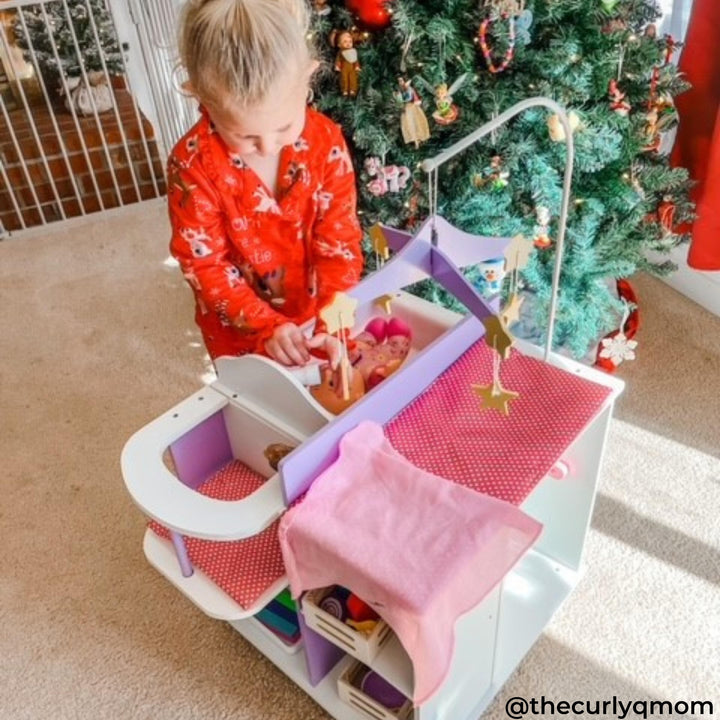 Child wearing red pajamas plays with a doll nursery playset near a Christmas tree.