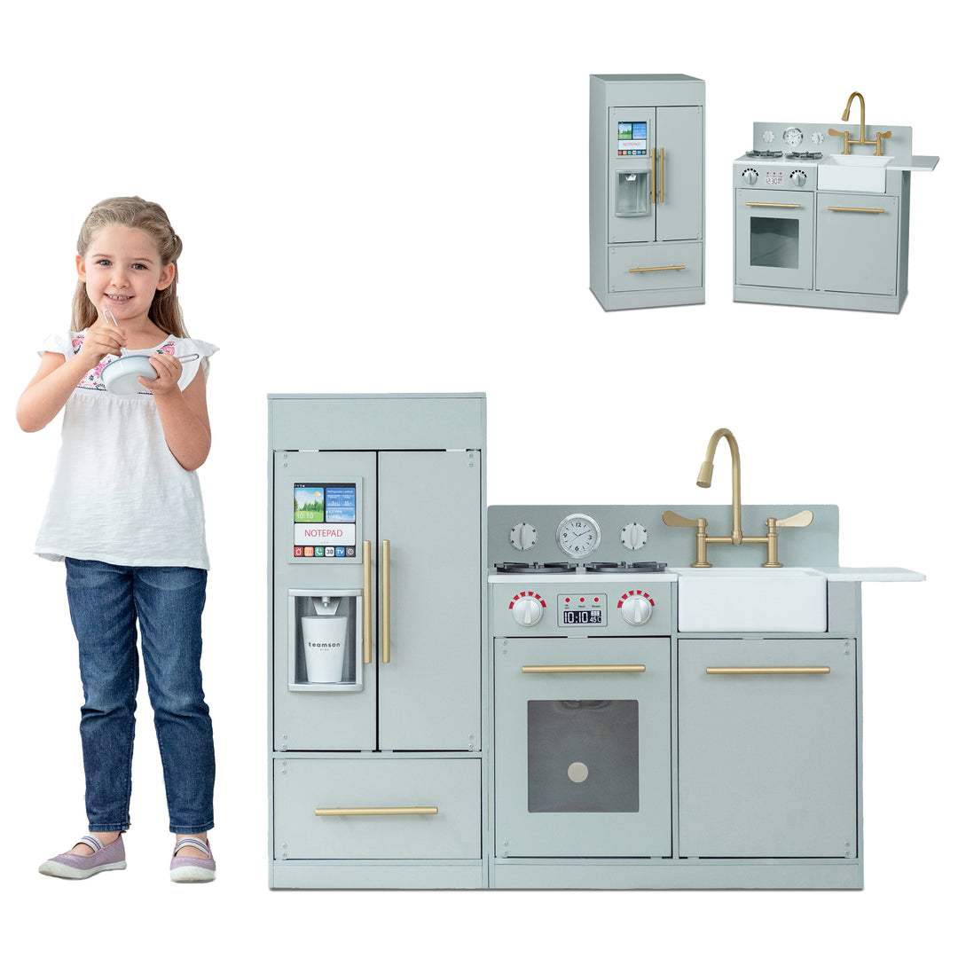 A young girl standing next to a Teamson Kids Little Chef Charlotte Modern Play Kitchen, Silver Gray/Gold, smiling and holding a teacup and saucer.