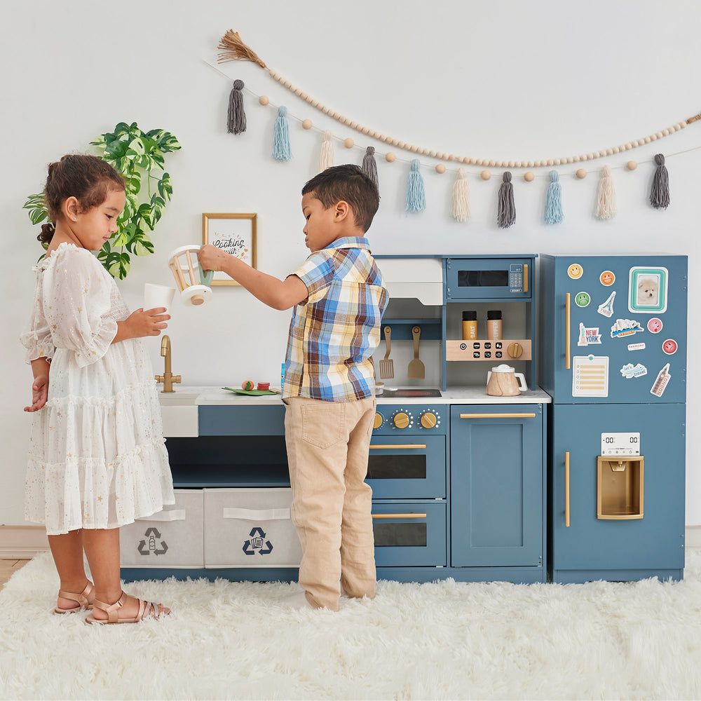Two children standing in front of a Teamson Kids - Little Chef Atlanta Large Modular Play Kitchen, Stone Blue/Gold.