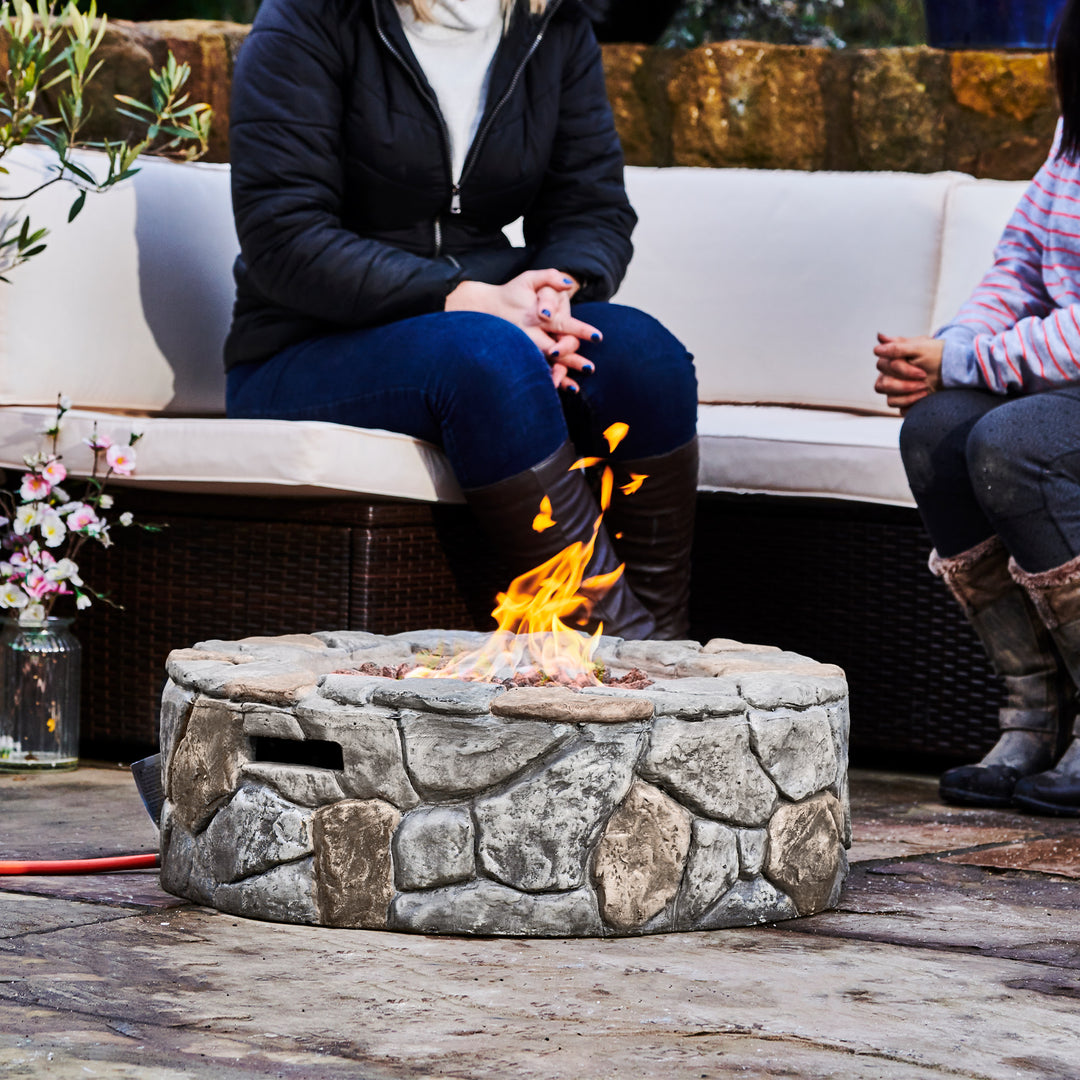 Two people sitting by a Teamson Home 28" Outdoor Round Stone Propane Gas Fire Pit, Stone Gray outdoors.