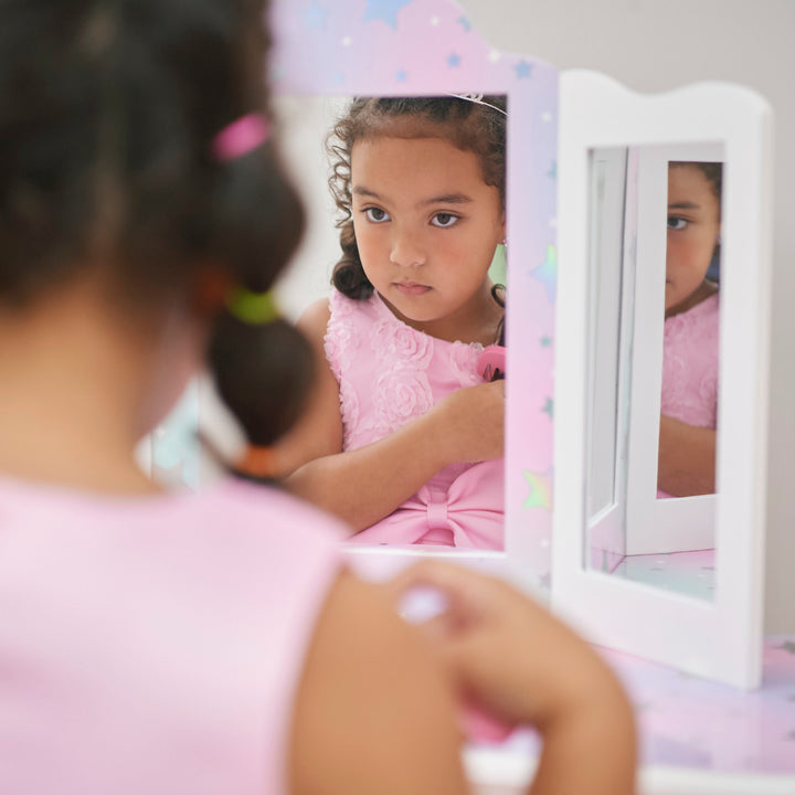 A little girl looking at herself in a the mirror of white vanity table with iridescent accents with silver stars.