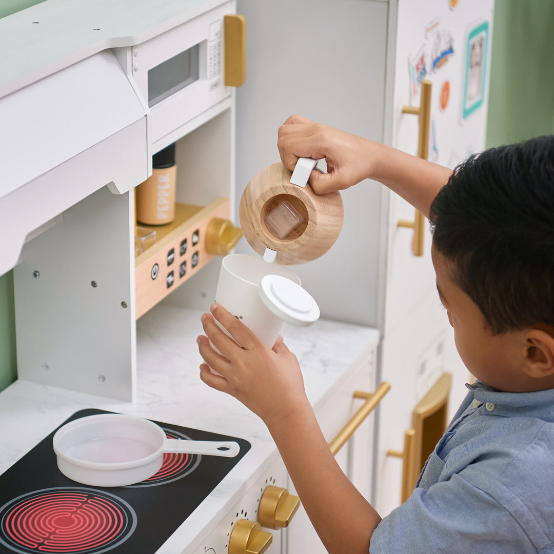 A child pretending to pour from a toy coffee pot into a cup at a Teamson Kids - Little Chef Atlanta Large Modular Play Kitchen, White/Gold station.