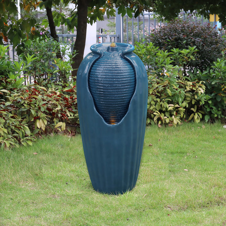 A large blue Teamson Home Indoor/Outdoor Contemporary Glazed Contoured Vase Water Fountain with LED Lights, now a water fountain, outdoors surrounded by greenery.