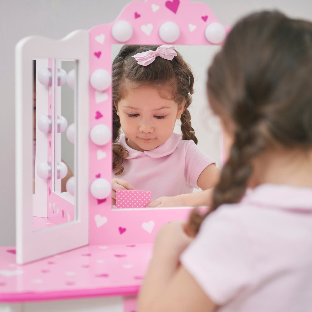 A little girl is sitting in front of a white and pink vanity set with table and stool with pink heart accents and a lighted tri-fold mirror.