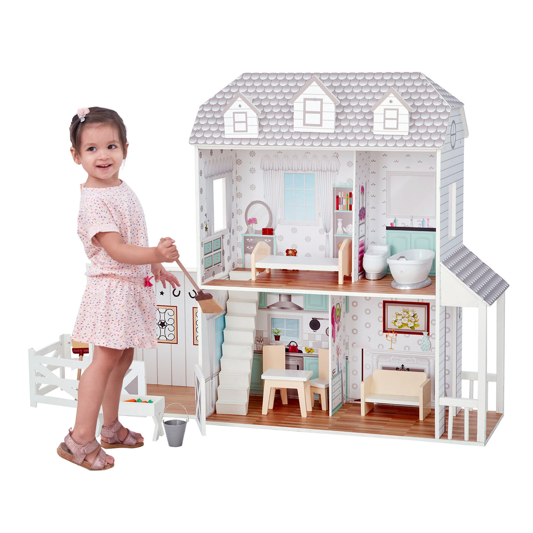 A little girl standing in front of the Teamson Kids Dreamland Farm Dollhouse with 14 Accessories, White/Gray holding a broom.