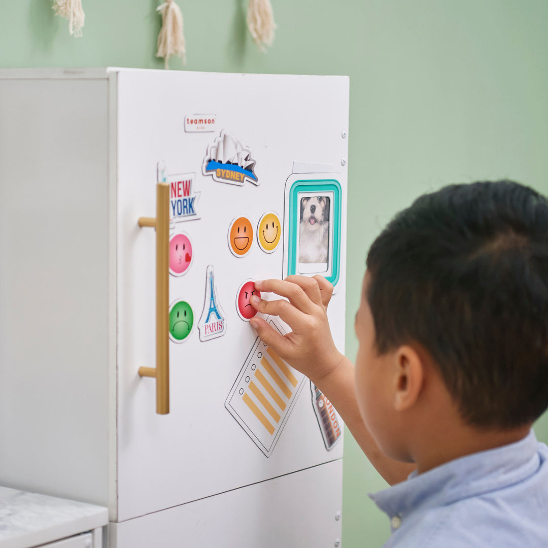 Child placing a TEAMSON KIDS - LITTLE CHEF ATLANTA LARGE MODULAR PLAY KITCHEN, WHITE/GOLD magnet on a refrigerator door decorated with various magnets.