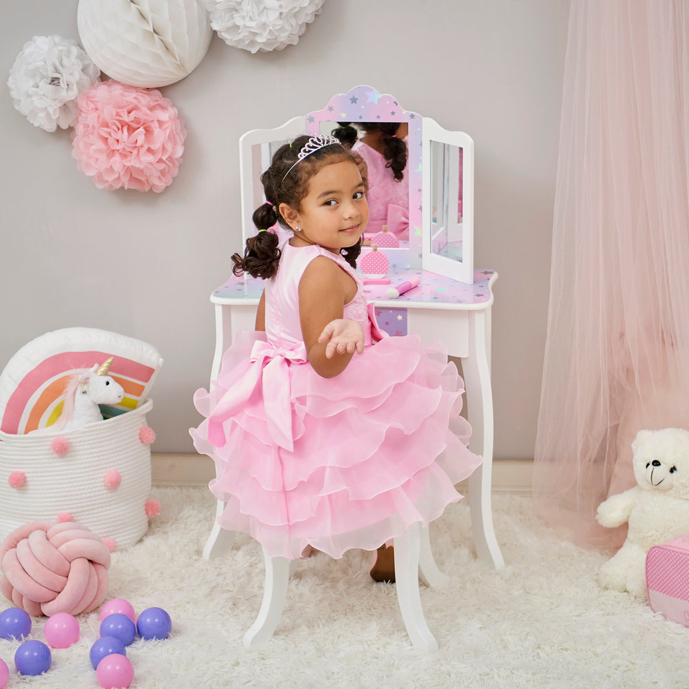 A little girl in a pink dress sitting in front of a Fantasy Fields - Gisele Starry Sky Print Vanity Set, White/Lavender mirror.