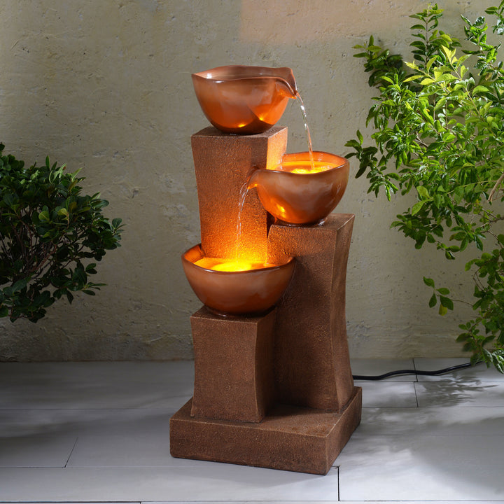 28.54" 3-Tier Outdoor Water Fountain with LED Lights, Brown with cascading bowls and led lighting in a darkened environment with plants on either side